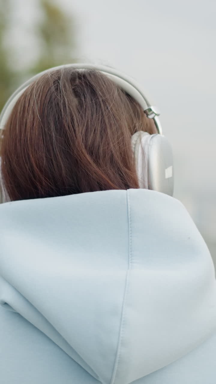 Real view of female artist in hoodie and headphones strolling, blurred withered tree and stainless rail in background, peaceful outdoor setting, casual walk, urban environment, relaxed mood
