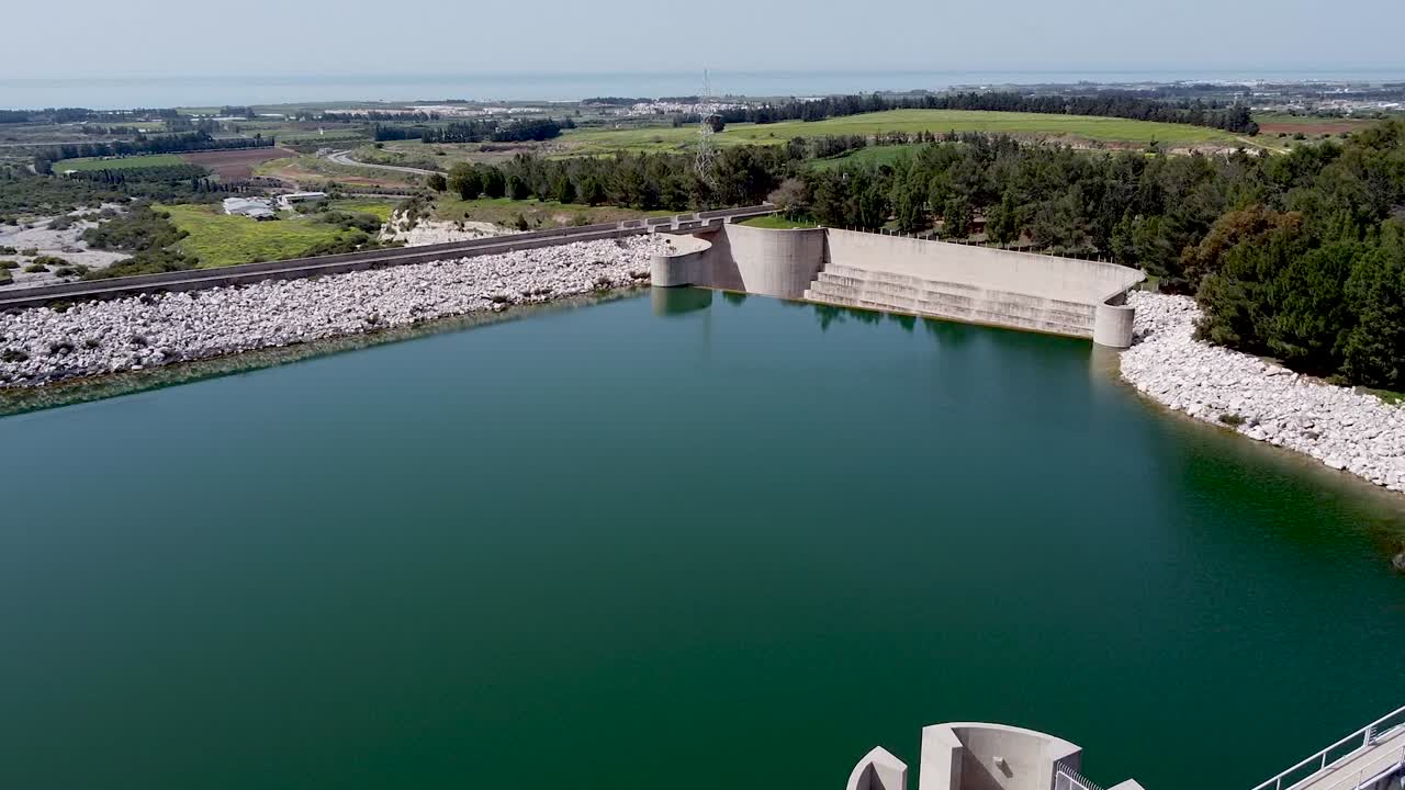 Observe the stunning aerial view of a tranquil reservoir featuring a distinctive tower. The surrounding green fields and clear skies enhance the peaceful atmosphere