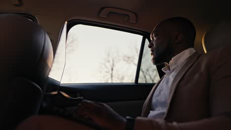 A-confident-man-with-Black-skin-and-a-beard-in-a-brown-jacket-works-on-a-laptop-and-looks-out-the-car-window-while-driving-a-modern-car-outside-the-city