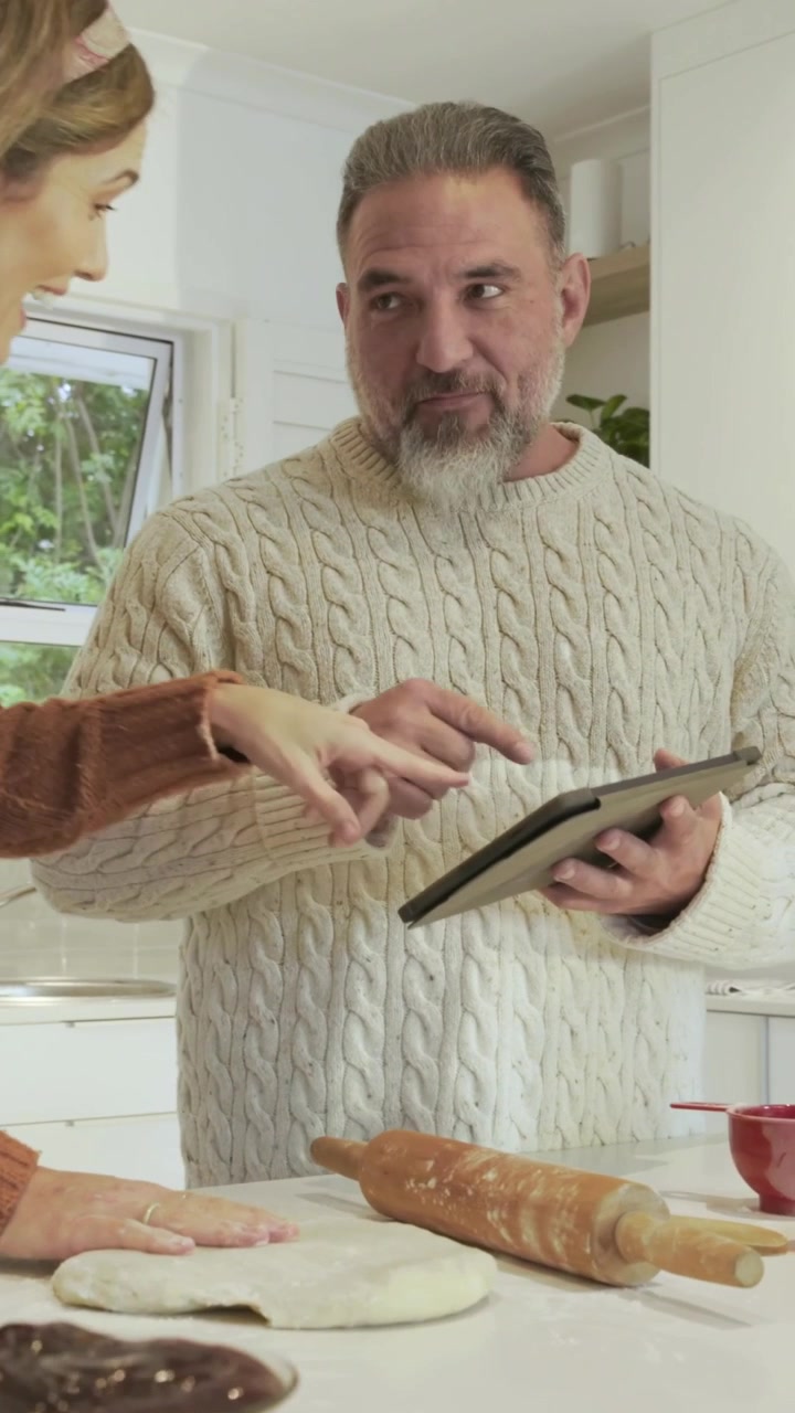 Vertical video: Tapping tablet guiding man in sweater through dough kneading, egg adding in kitchen
