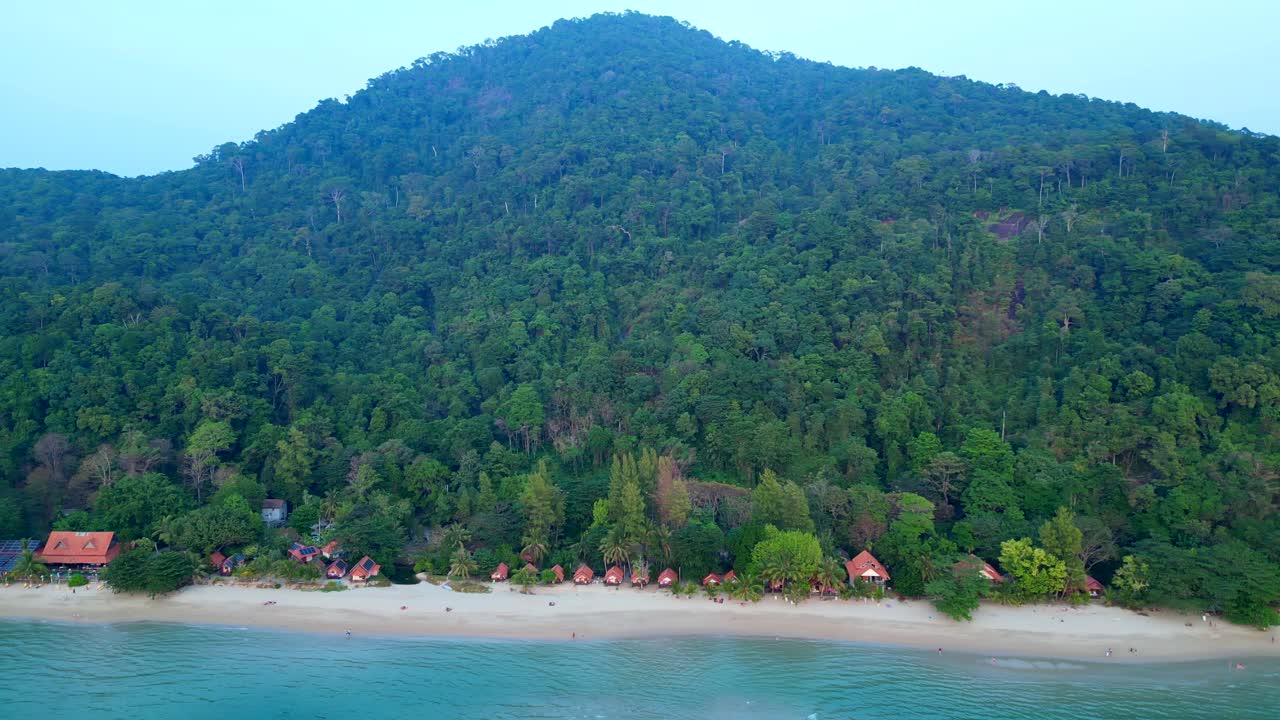 Koh Chang Tropical beach with bungalows along the shore and dense green jungle covering a mountain. Brilliant aerial view dolly left panorama overview drone
