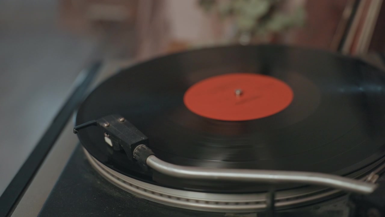 Primo piano di un disco in vinile su giradischi, atmosfera calda e vintage con etichetta rossa, puntina che traccia i solchi, luce ambientale e leggero fruscio, rituale da collezionista in un accogliente soggiorno che suggerisce un ascolto da audiofilo.