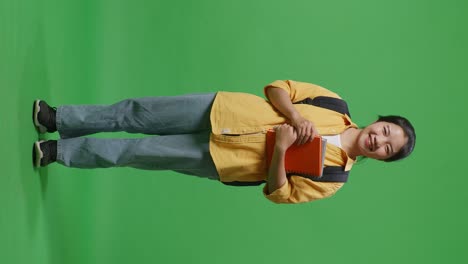 full body of asian woman student with a backpack and some books standing and smiling to camera in the green screen background studio