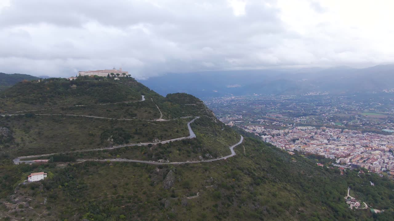 Scenic view of abbey of Montecassino on summit of green rocky hill with downtown Cassino city and buildings beneath on cloudy day, Italy, sideways aerial