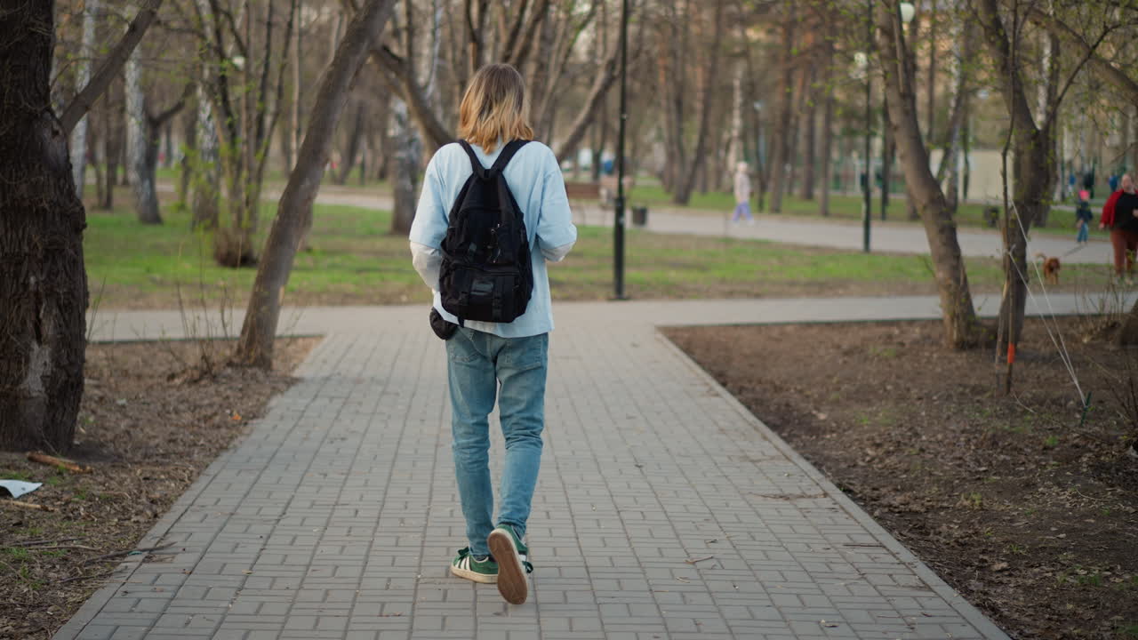도시 공원 포장 도로를 따라 걷는 스케이트보더, 어깨에는 백팩, 데님 청바지, 포장 도로를 치는 스니커즈, 뒷모습 모션, 꾸준한 속도, 자연광, 산책로를 따라 늘어선 나무, 도시
