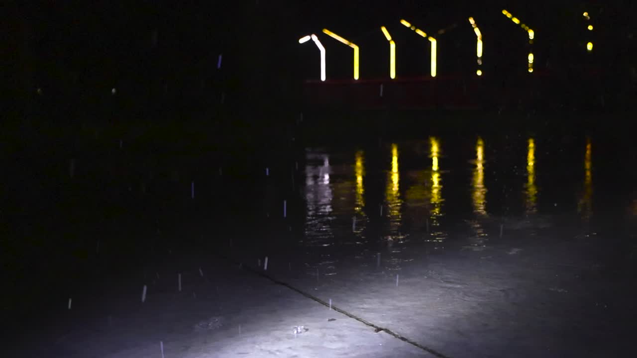 Gorgeous and soothing slow motion footage of raindrops falling on a water covered concrete slab in a skatepark during a stormy evening or night and street light reflecting on water in the bokeh back