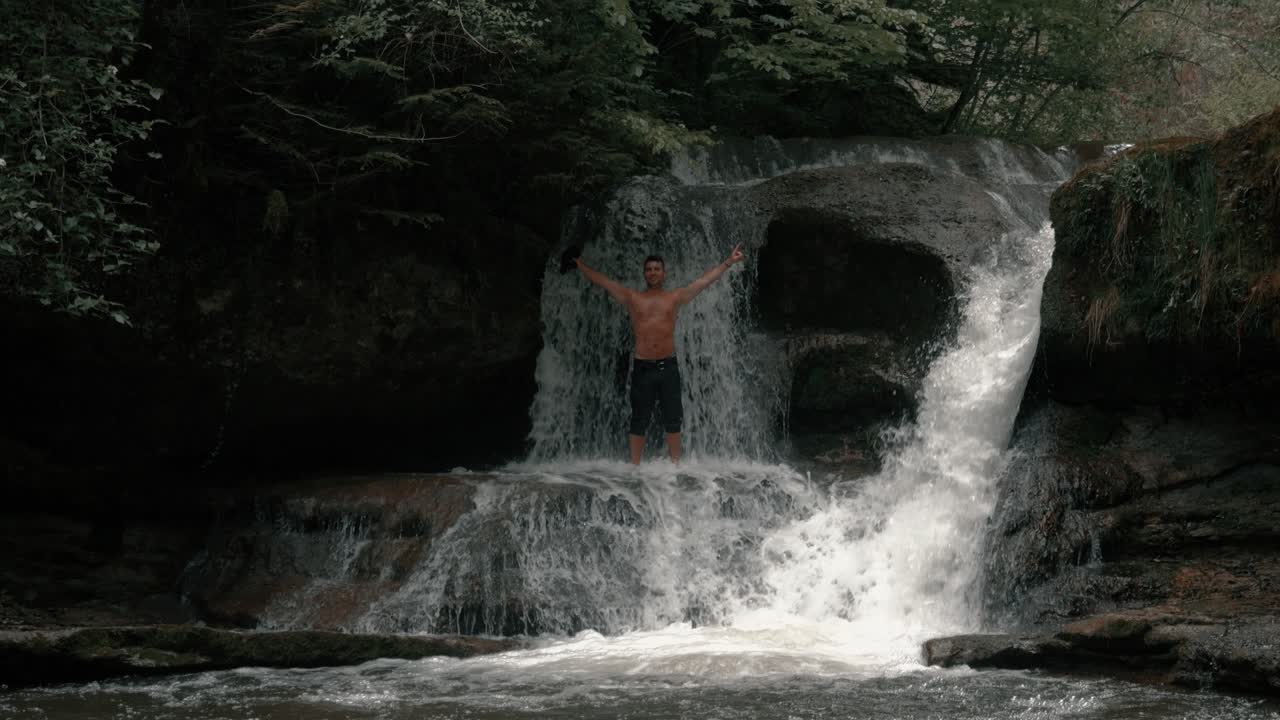 A man stands in front of a waterfall in an epic forest. It's mystical, and the man is shirtless. He stretches his arms in the air, enjoying nature. Shot in slow motion