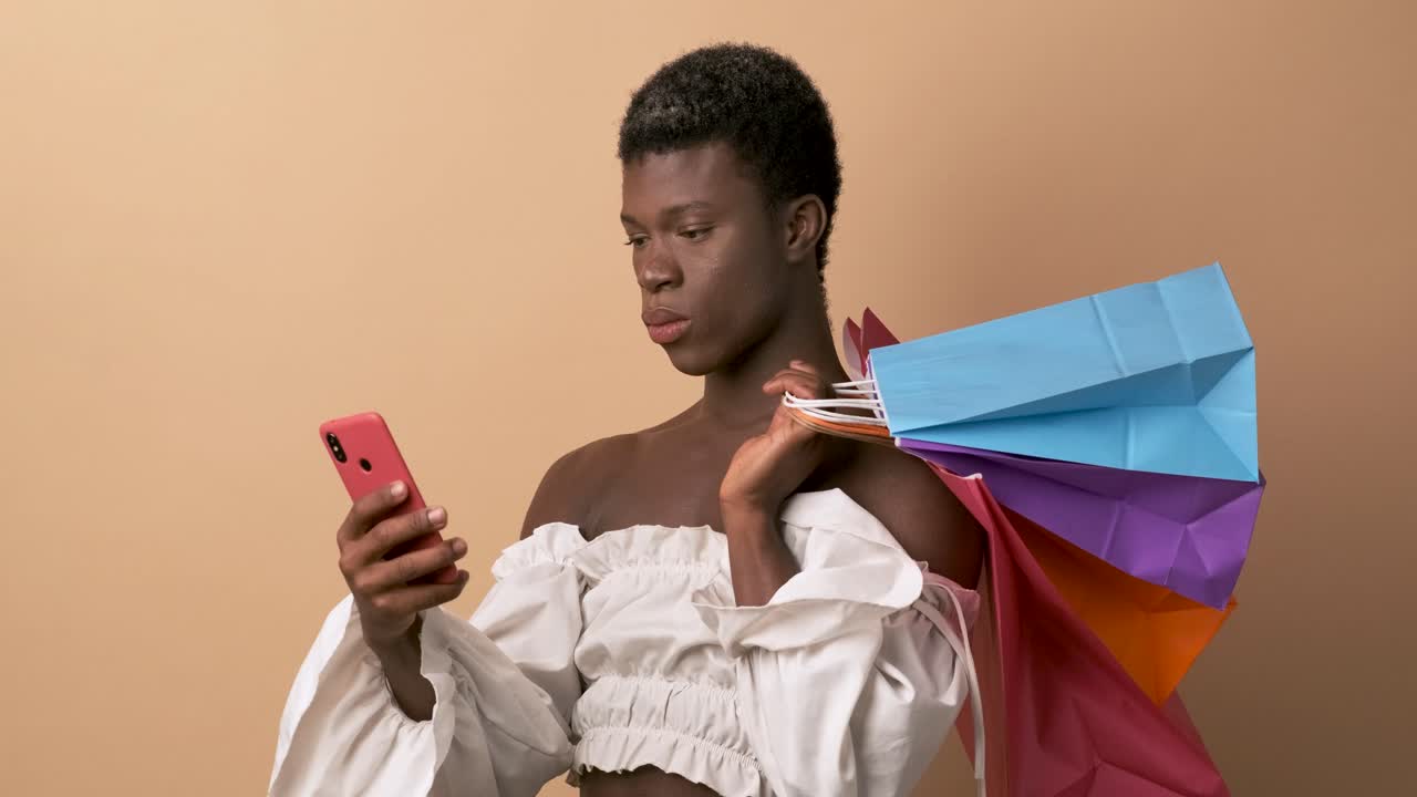 Transgender african person using phone and holding shopping bags over beige background