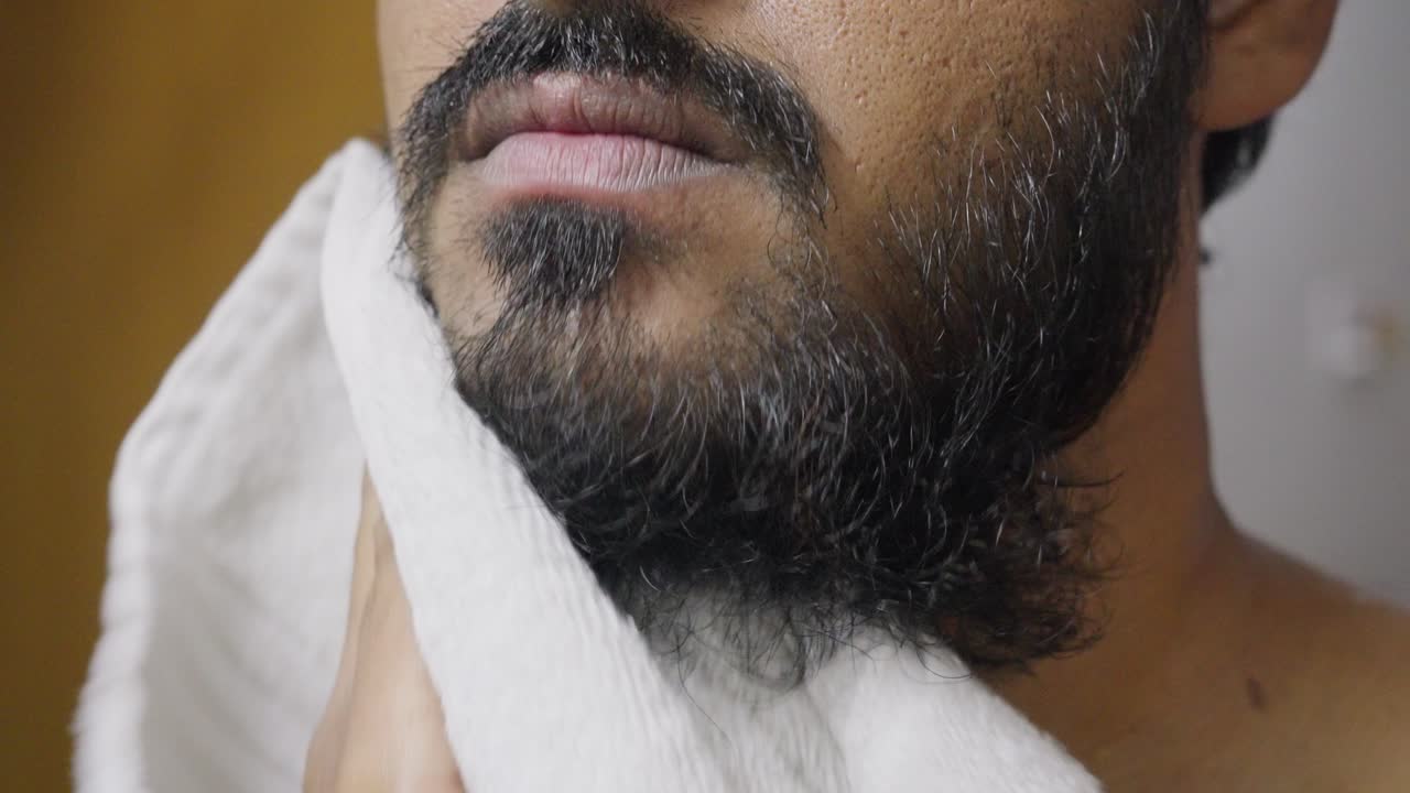 A closeup footage of a young man drying his beard with a white towel, with blurred background. Hygiene concept