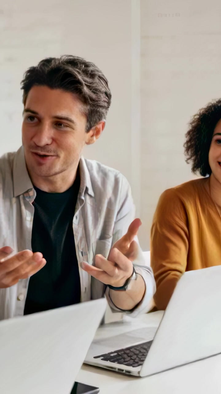 Vertical video: Starting man in gray shirt gesturing and making point at table with laptops phone