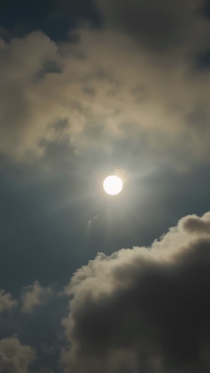 Vertical video: Drifting wispy and cumulus clouds causing light shifts in sun disc in open sky
