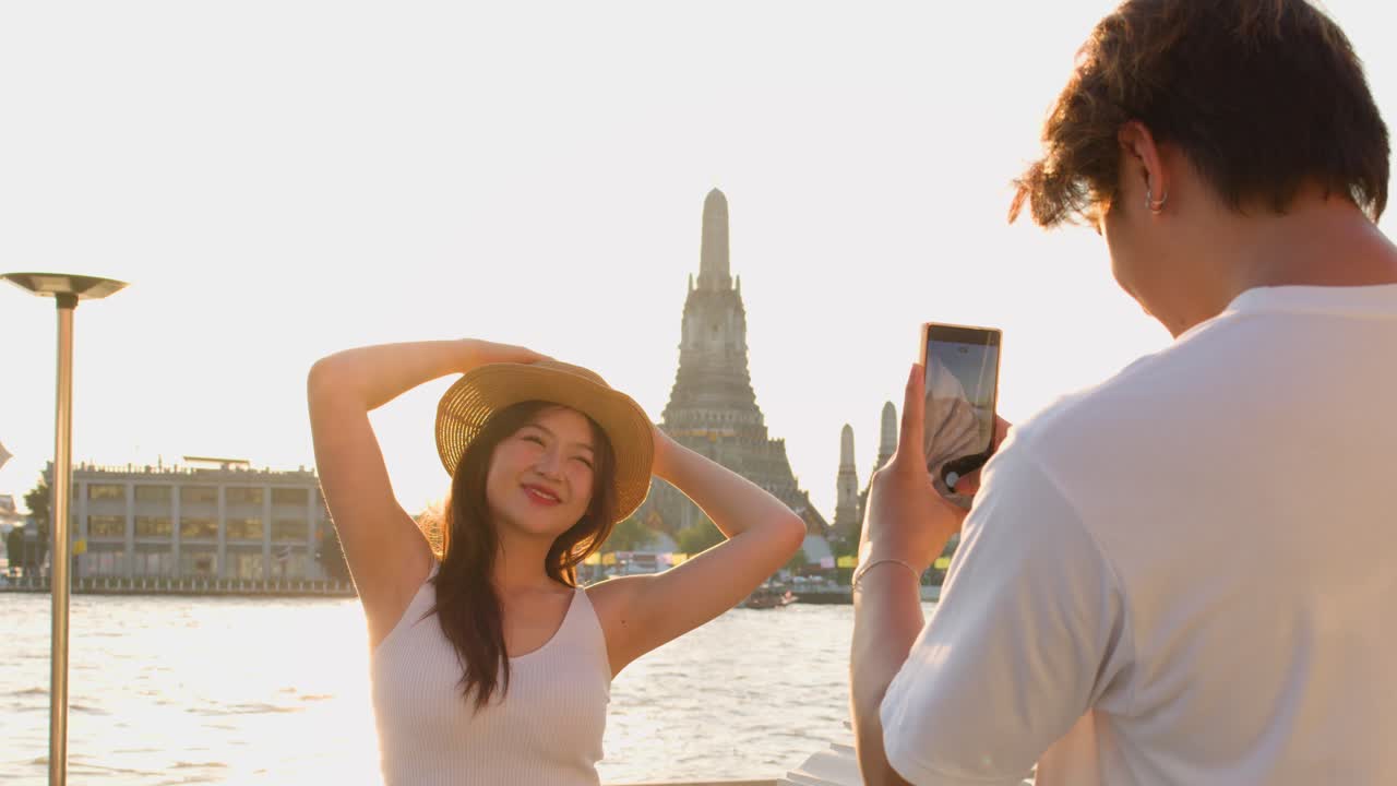 Donna sorridente posa giocosamente mentre un uomo la fotografa vicino a un tempio sul fiume nella calda luce del tramonto