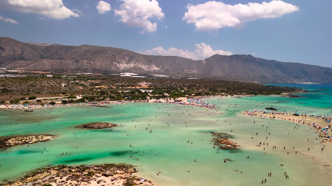 Visitors enjoy a sunny day at Elafonissi Beach in Crete, surrounded by stunning turquoise waters and scenic mountains. The shore is filled with happy beachgoers and sunbathers
