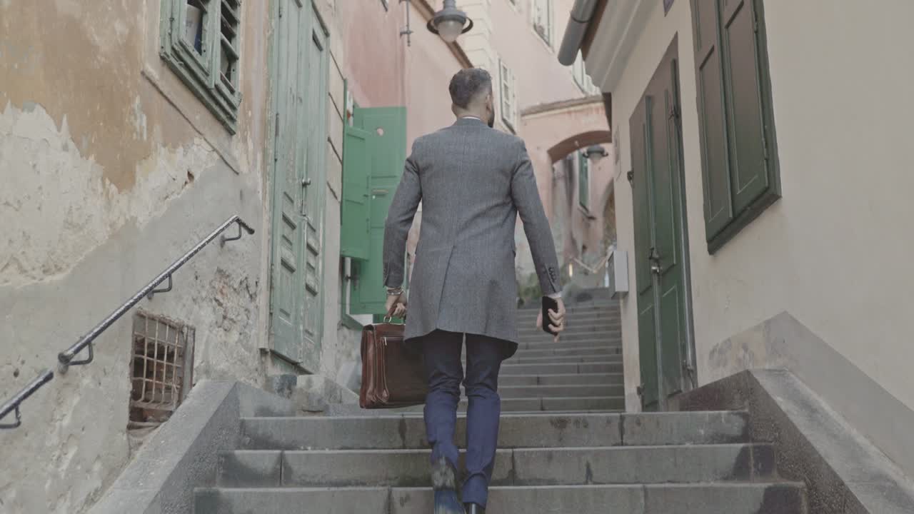 vista trasera de un elegante joven en traje con maletín y abrigo largo subiendo las escaleras y mirando hacia el lado