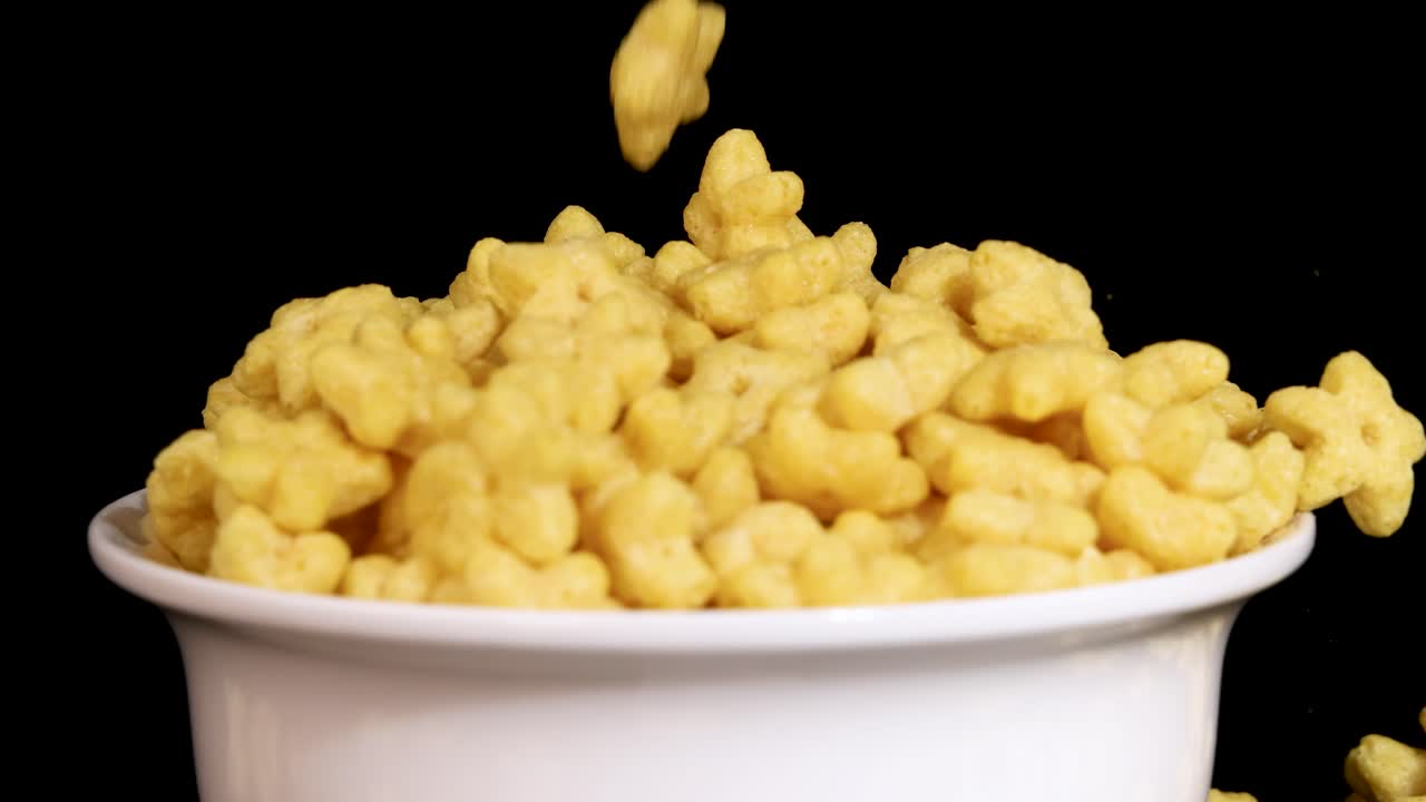 Yellow cereal pieces fall into a white bowl against a black background, captured in slow motion with clear lighting