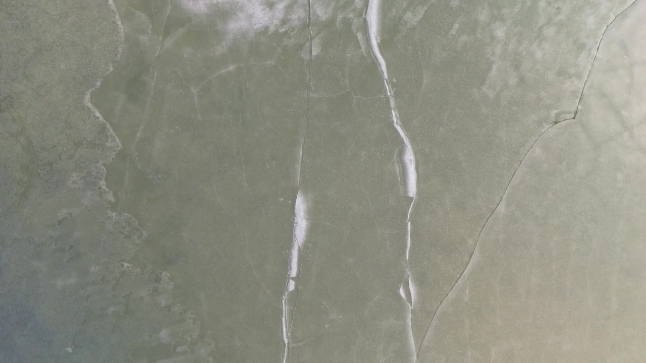At Orava Reservoir in northern Slovakia, the frozen lake displays intricate ice cracks and natural patterns within a quiet winter landscape, seen from a tilt-down drone view.