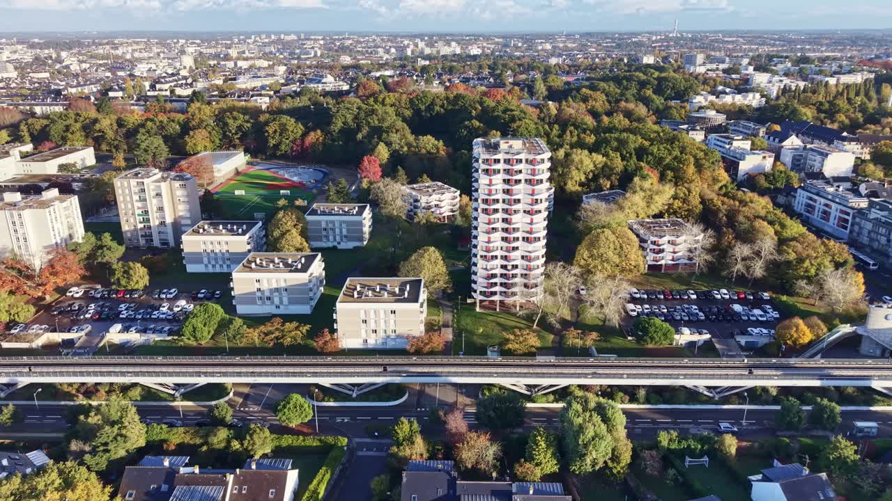 Drone panning shot above La Poterie metro station in Rennes, moving left to right while following the metro line as a train passes in the opposite direction under the sunrise light