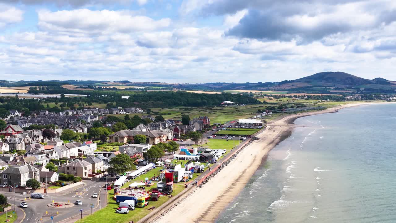 Drone glides above Fife coastline, revealing seaside town, sandy beach, and distant hills in daylight