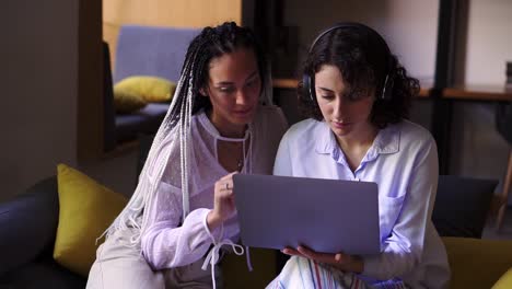 Two-stylish,-focused-female-friends-sitting-on-grey-and-yellow-couch-with-one-laptop,-studying-or-browsing-smth-together.-Two-students-working-remotly-with-one-laptop.-Modern-interior.-Close-up