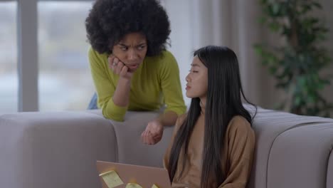 upset frustrated asian woman sighing looking at laptop screen supported by african american lady. sad young depressed freelancers working remotely in home office living room.