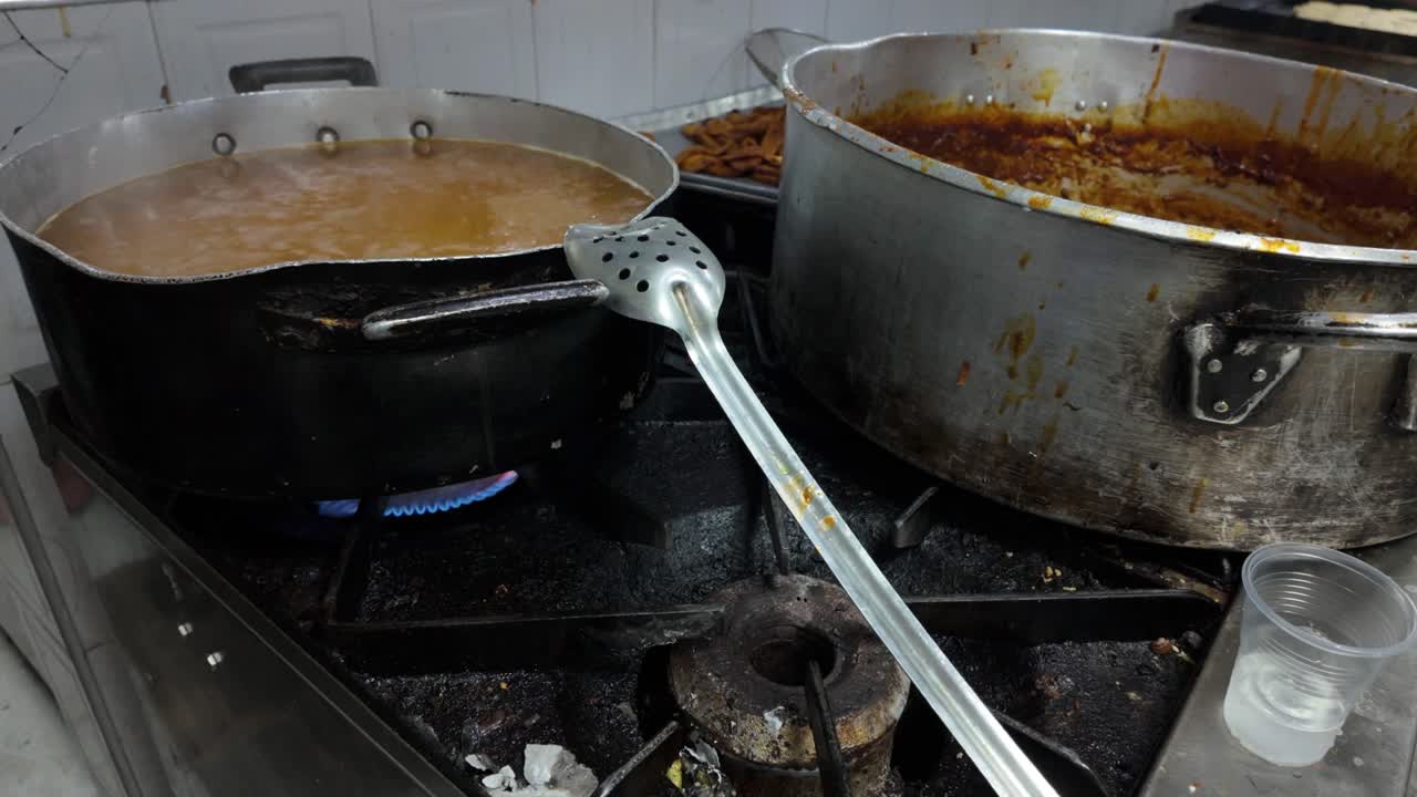 Tilt down documenting the large-volume preparation of Latin American staples like *carne mechada* (shredded meat) and beans, on an industrial stove inside a bustling commercial kitchen