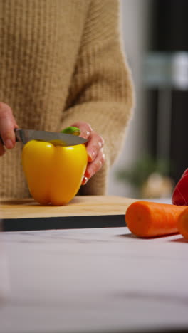 vertical video close up of woman at home in kitchen preparing healthy fresh vegetables for vegetarian or vegan meal slicing yellow peppers on board