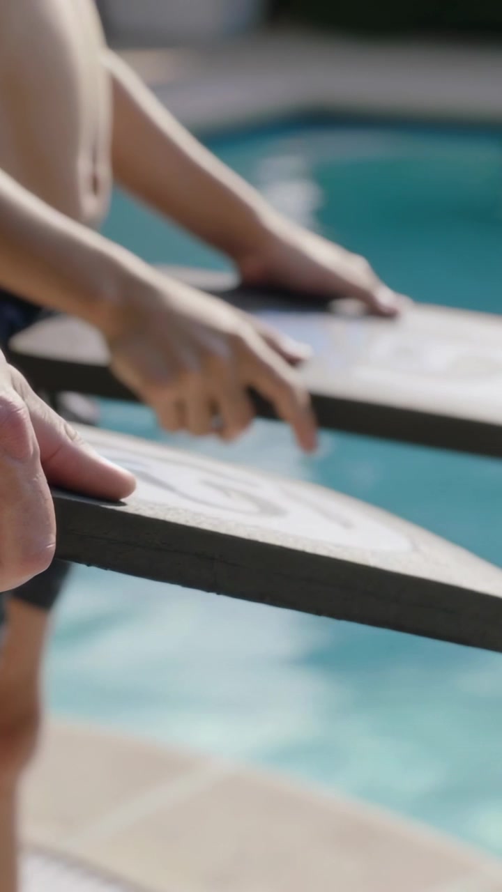 Vertical video: Gripping ledges, two swimmers adjusting fingers, leaning at pool edge in trunks