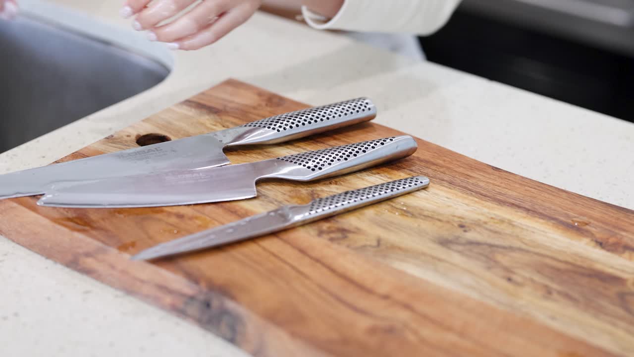 A person arranges knives on a wooden cutting board in a well-lit kitchen setting