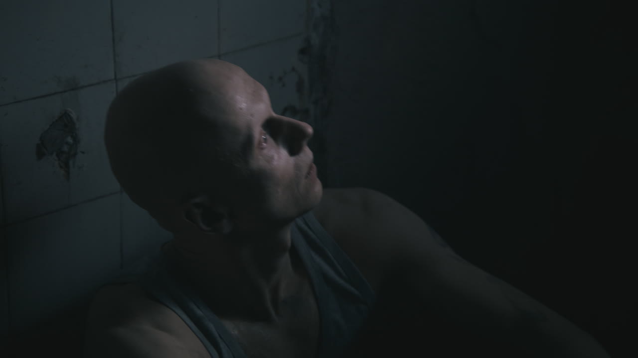 Un uomo pallido, calvo e magro si siede accanto al muro nel vecchio bagno inquietante di notte.