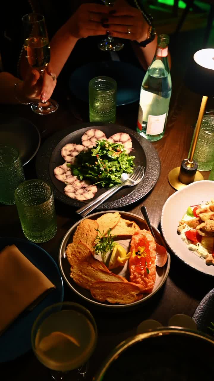 Overhead view of seafood appetizers and wine at dinner. Restaurant table setting with octopus carpaccio, salmon tartare, toasted bread and green glasses in warm light
