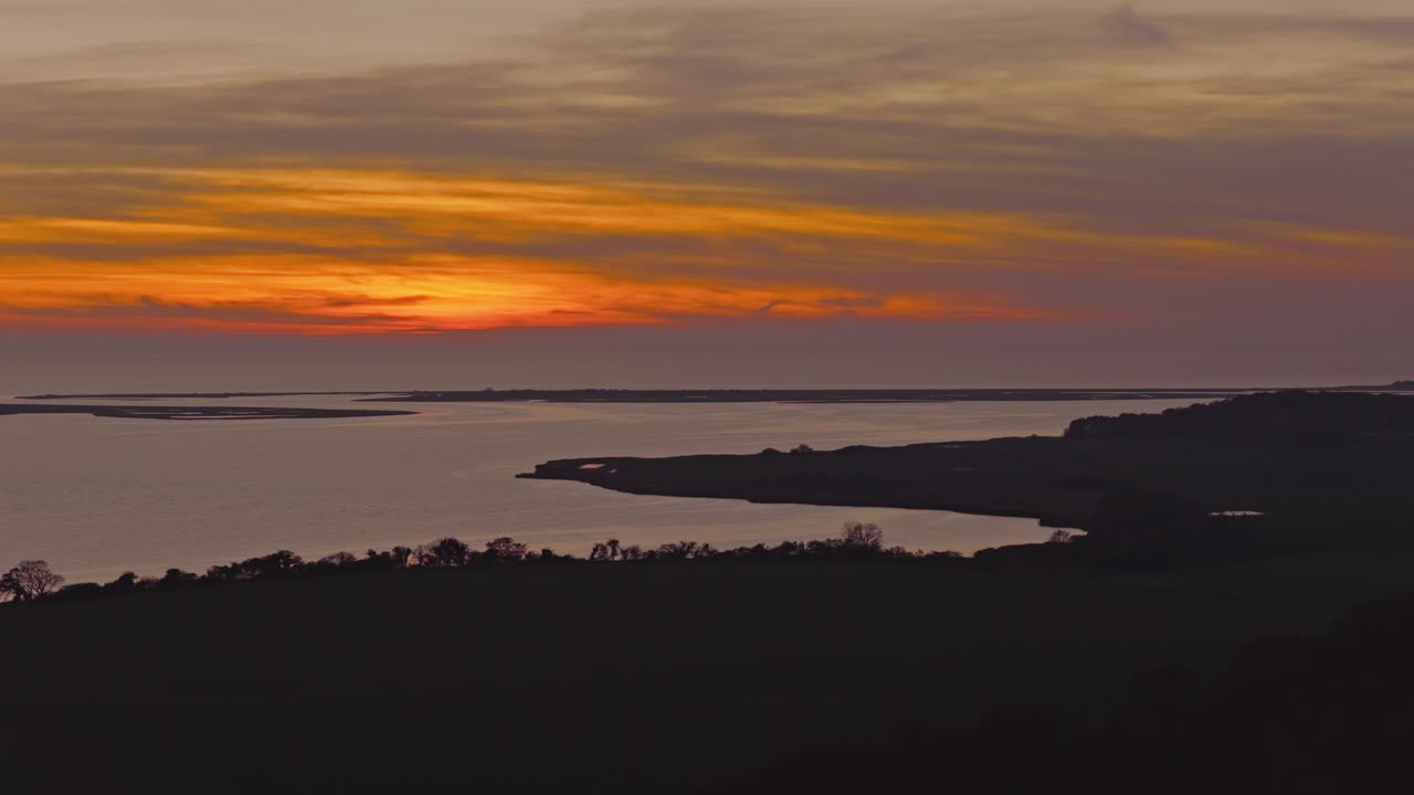 Warm golden and orange hues illuminate the evening sky above a calm coastal inlet of a scandinavian fjord. Silhouetted landforms and gentle water reflections create a peaceful nordic sunset mood