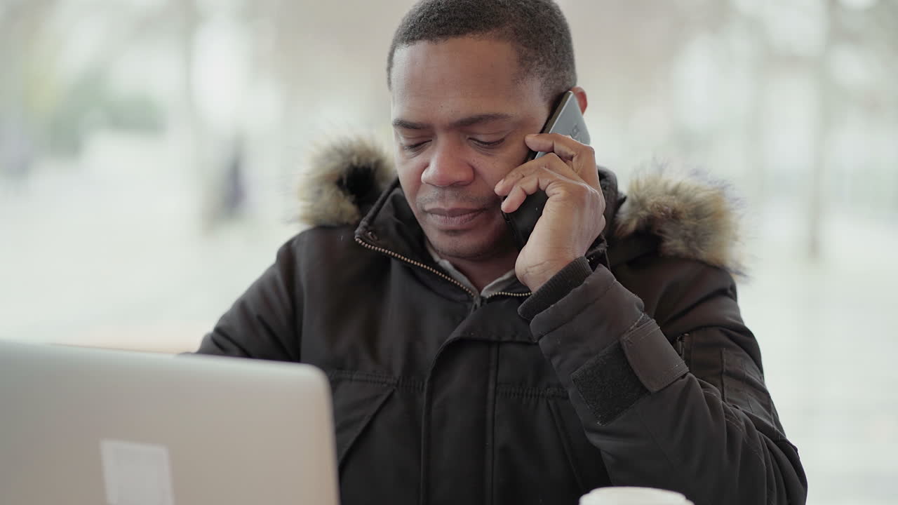 Afro-American middle-aged man in black jacket with fur hood working on laptop outside