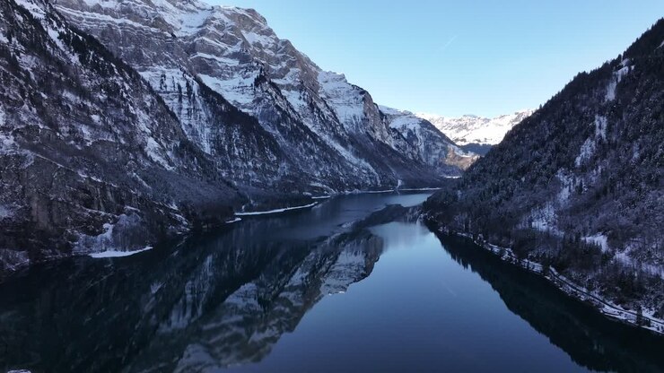 Aerial view of Klöntalersee in Switzerland reveals the lake winding like a ribbon amidst majestic, snow-covered mountains. The still water perfectly mirrors the towering peaks, enhancing the scene.