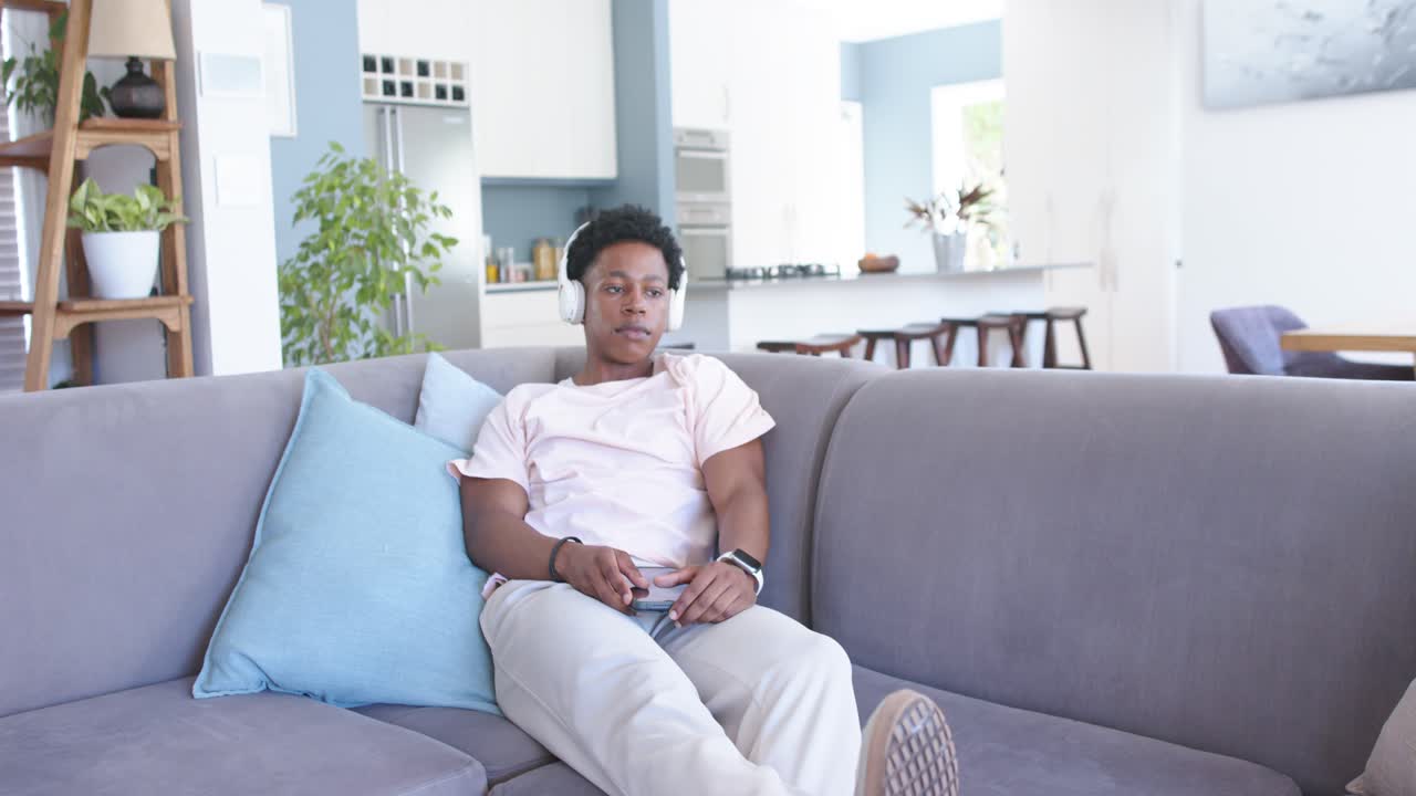 African American man sitting on gray couch at home, using white headphones and phone for relaxing