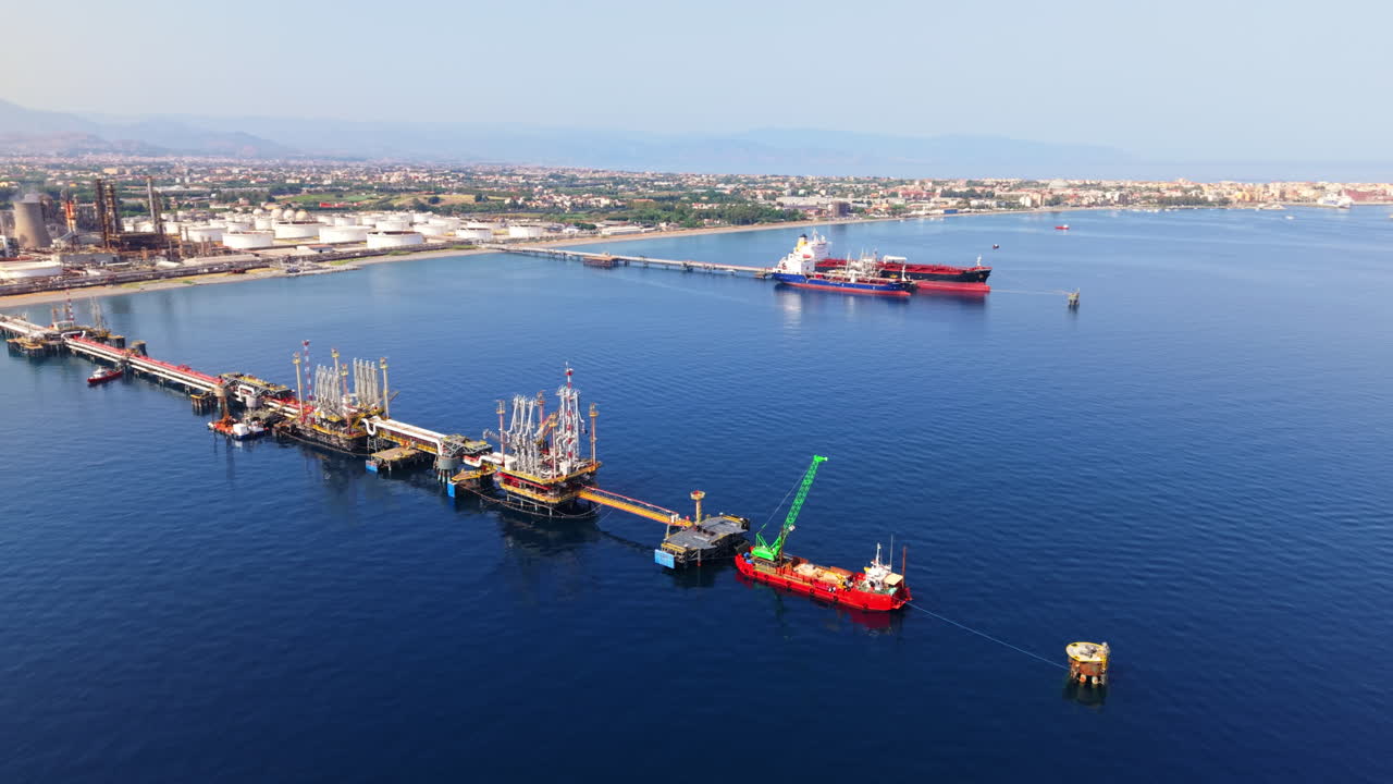 Drone shot descending with tilt over offshore oil station linked to refinery on Sicilian coast, with tankers moored at piers