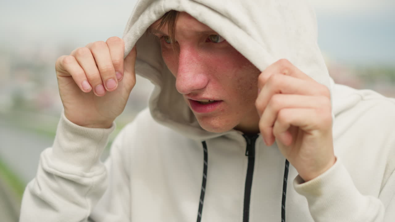 비를 흠뻑 맞은 후드티를 입고 머리카락이 젖어 이마에 붙고, 후드를 아래로 잡아당기고, 감정이 북받쳐 눈을 감고 있는 젊은 백인 남성, 흐릿한 도시 스카이라인, 옷감에 맺힌 빗방울, 상처받기 쉽고 감정을 정화하는 모습