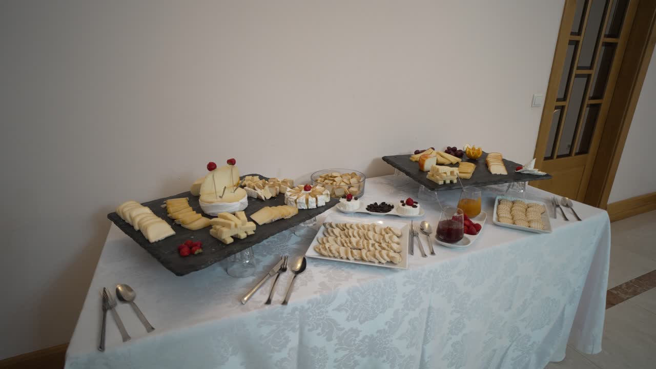 Elegant cheese and fruit buffet setup on a white tablecloth with various platters and utensils