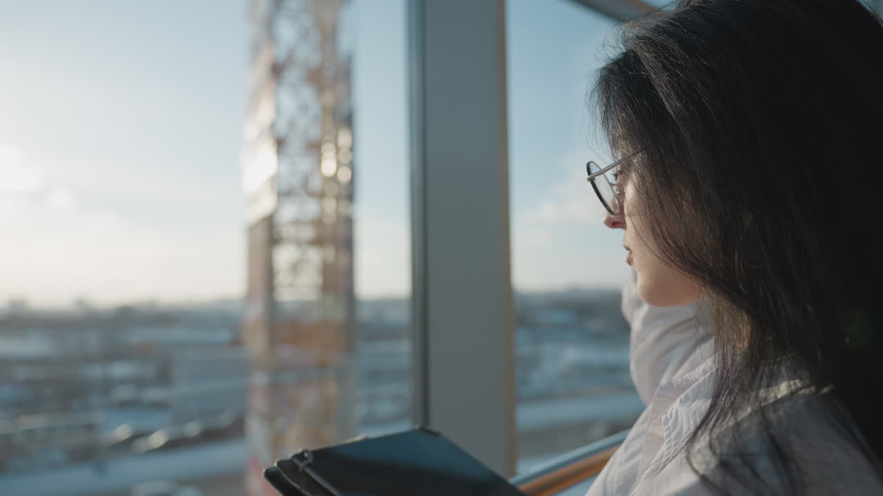 Side view of woman wearing glasses using tablet near large glass window with sunlight streaming in, her hand resting on her head while immersed in screen, cityscape visible through glass