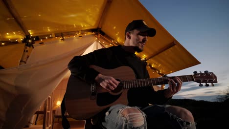 man plays the guitar and sings songs in a tent with light bulbs