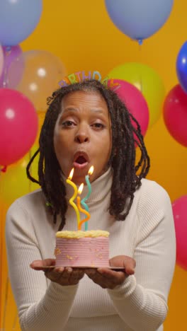 vertical video studio portrait of woman wearing birthday headband celebrating birthday blowing out candles on cake with confetti