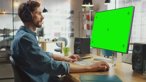 un joven empleado creativo profesional trabaja en su computadora personal con una gran pantalla verde. trabaja en un cool loft de oficina. un hombre creativo se pone auriculares.