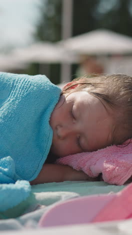 tired little girl with wet hair covered with towel sleeps on deck chair on beach at marine resort on sunny summer day close view slow motion