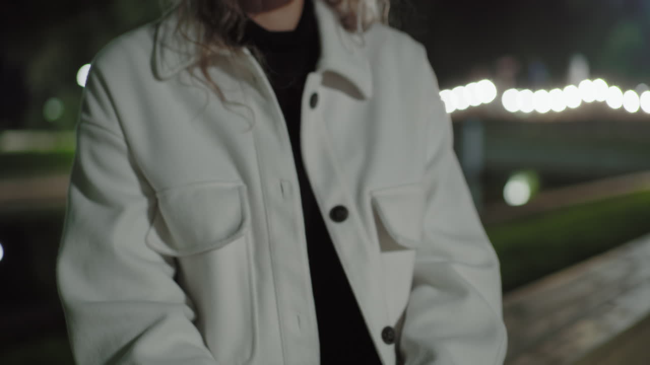 Woman seated near water holding book on her leg, lost in thought, with soft reflections of glowing lights on water surface behind her, moody nighttime atmosphere
