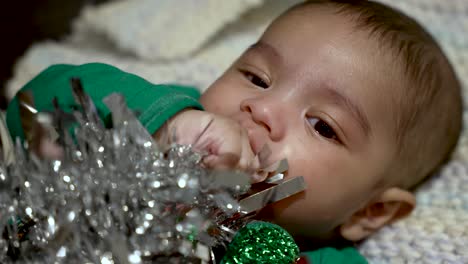 adorable 2 month bangladeshi baby boy lying on blanket sucking hand whilst grabbing silver tinsel