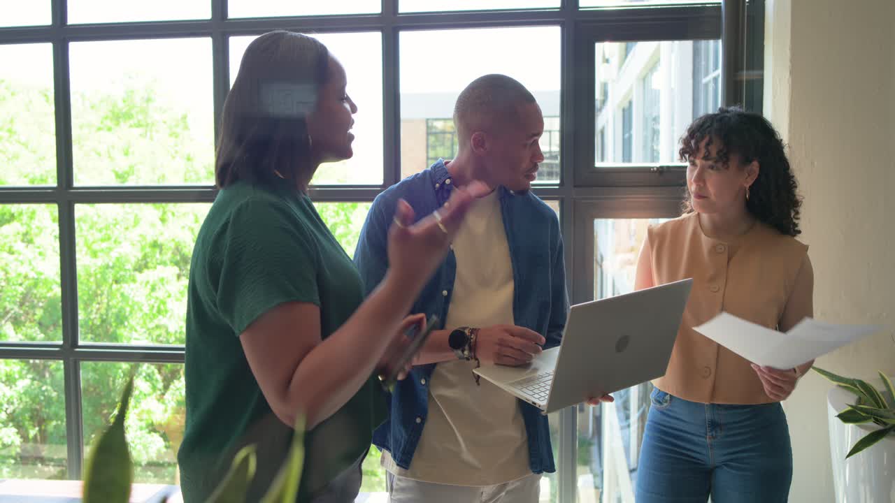 African American coworkers by window, man angling laptop while woman reading papers, reviewing work