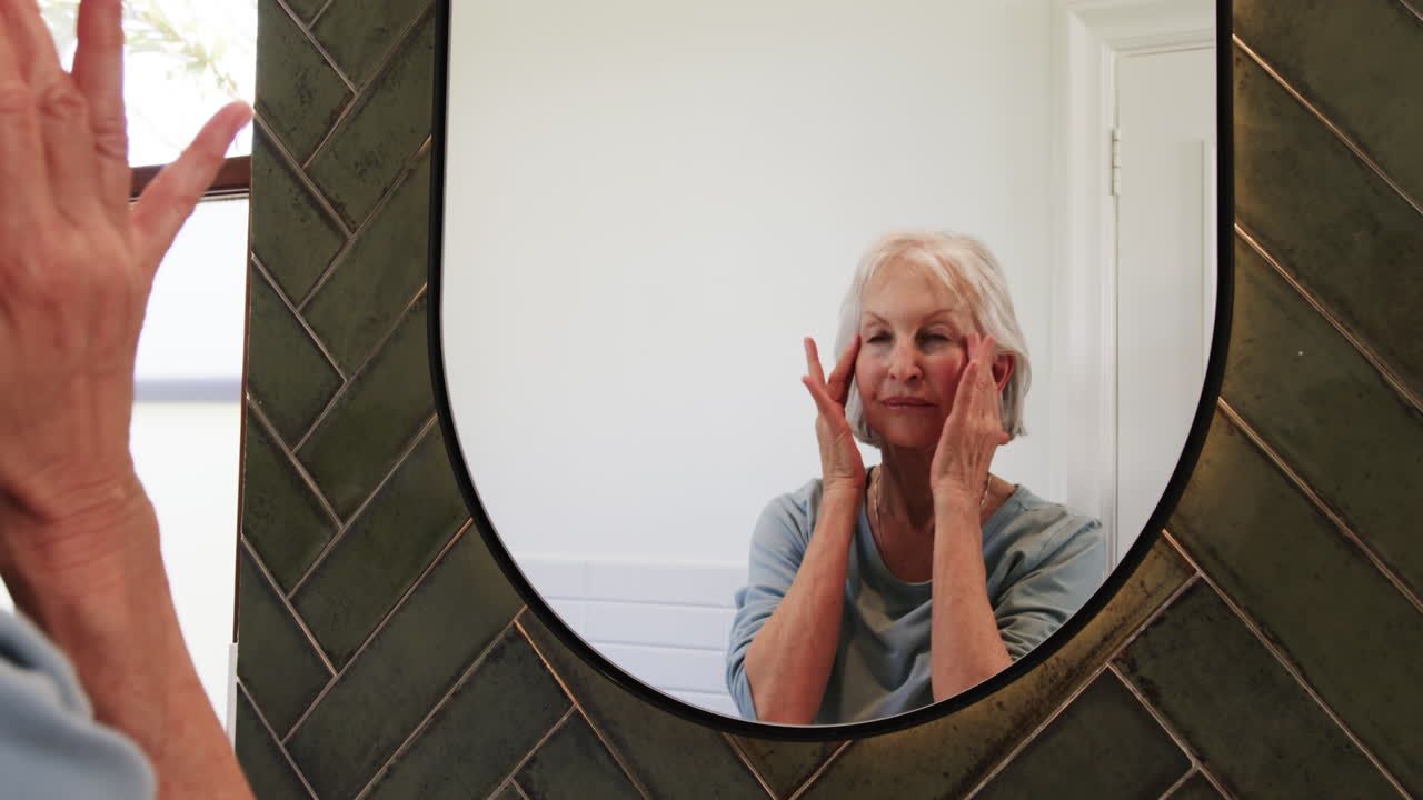 Senior woman gently massaging face while looking in bathroom mirror, at home