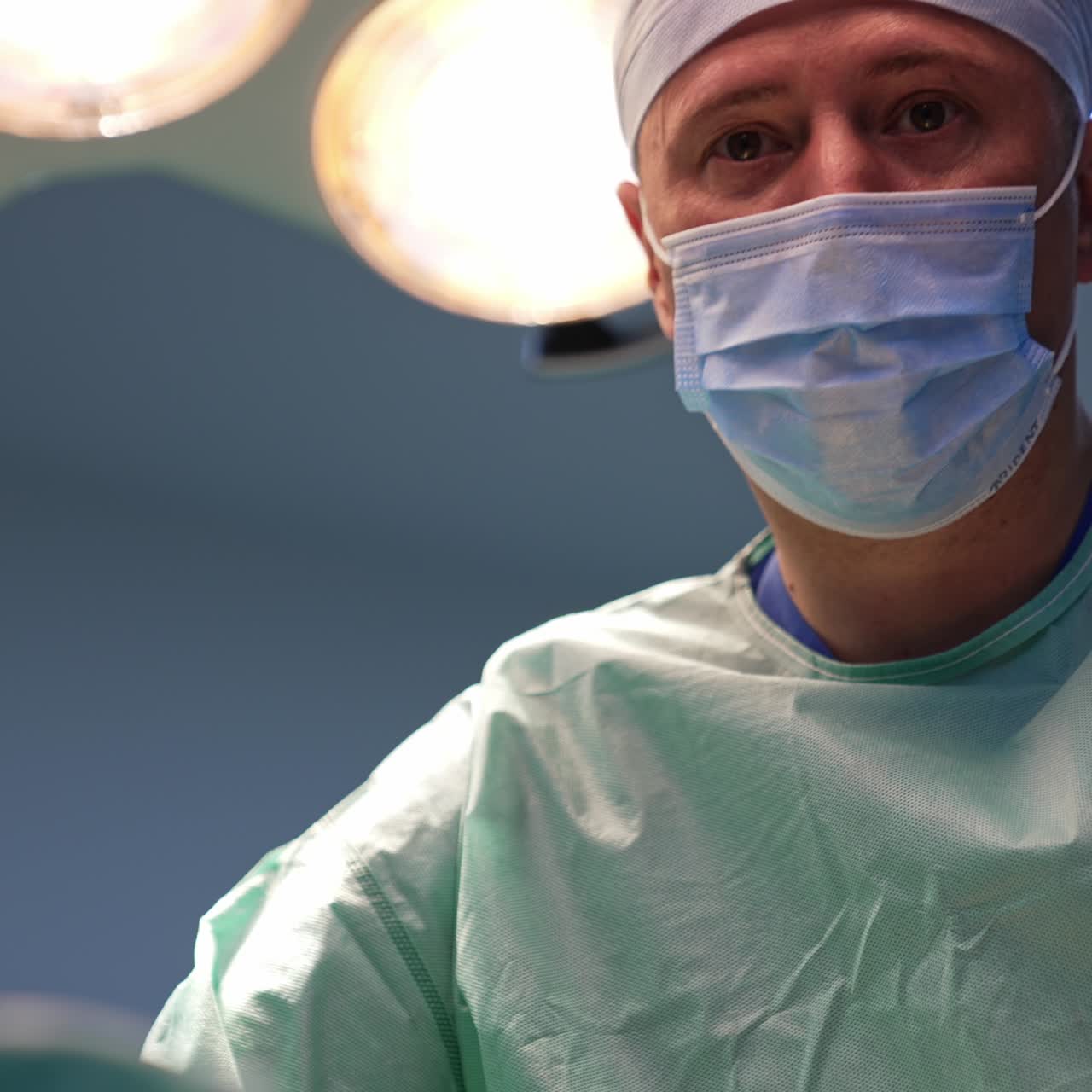 Doctor in white cap and mask bent over the patient under operation. Portrait of a confident surgery professional focused on his work
