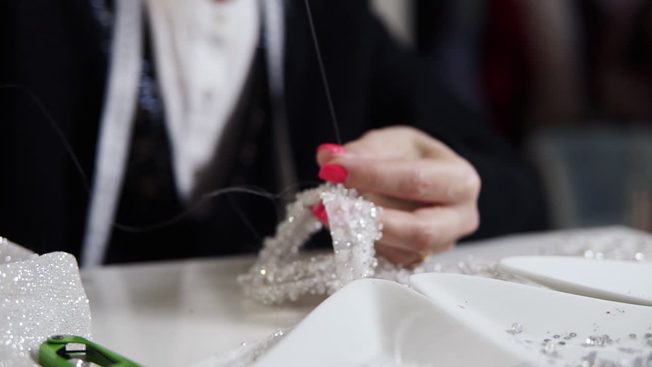 분홍색 매니큐어와 함께 여성 손의 클로즈업 바늘과 실을 들고 장식 돌로 독점적인 웨딩 드레스를 장식합니다. 우아한 웨딩 드레스를 바느질합니다. 손으로 만든