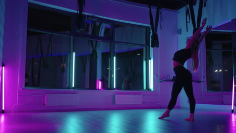 A-woman-does-yoga-and-stretching-on-a-hanging-hammock-in-neon-light.-A-woman-is-flying-in-a-hammock-in-the-studio-doing-stretching-and-body-exercises.-Exercises-in-the-air