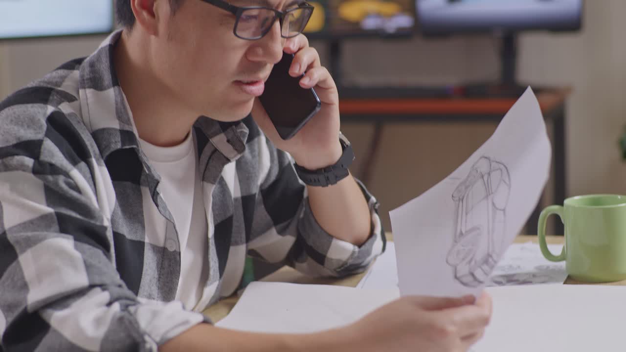 아시안 남성이 자신의 자동차 디자인을 손으로 스케치하고 스마트폰으로 이야기하면서 스튜디오에서 테이블 위에 자동차 디자인 스케치를 작업하고 있습니다.